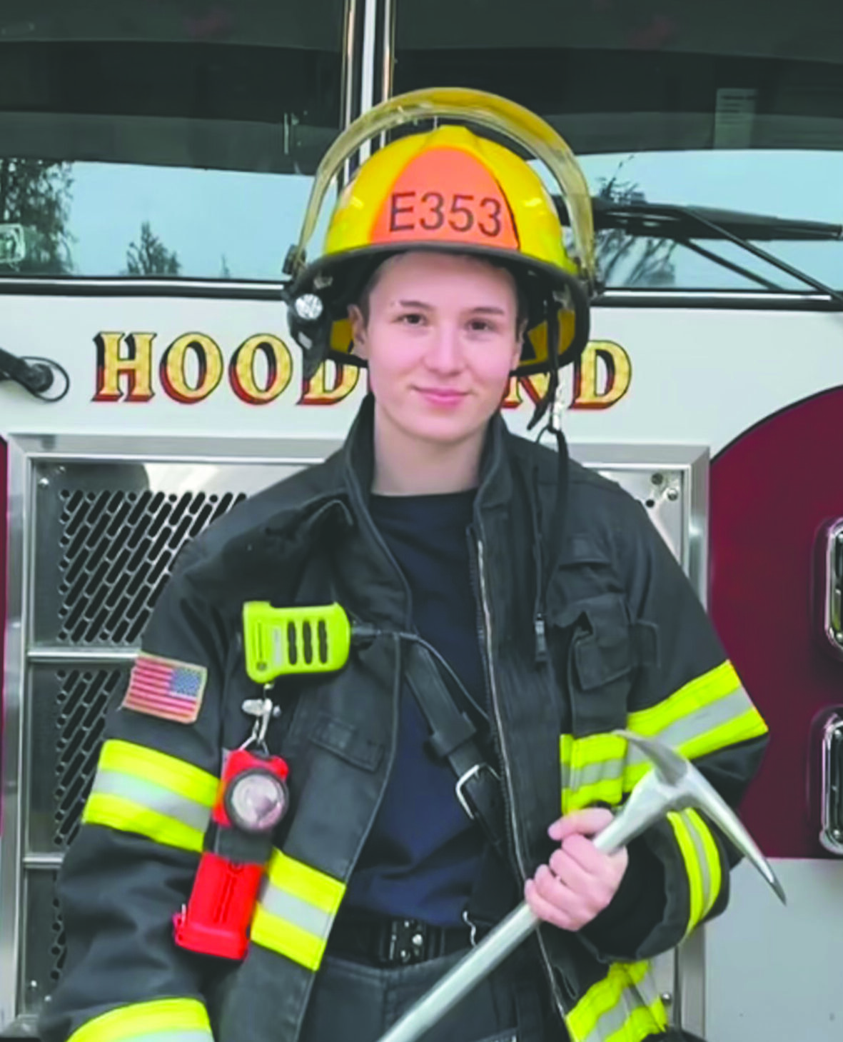 Fire fighter Kiera stands in front of a fire truck in full gear with her axe at the ready