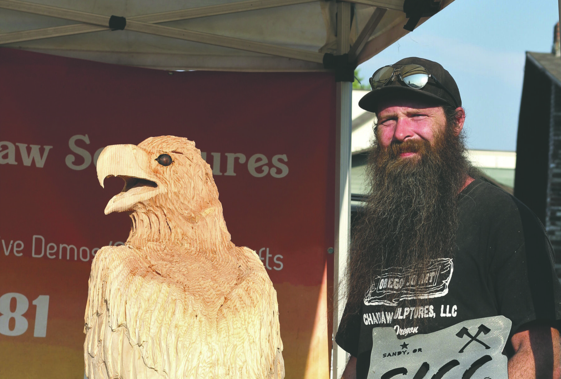 An artist stands in front of his chainsaw sculpture of an eagle, holding his trophy.