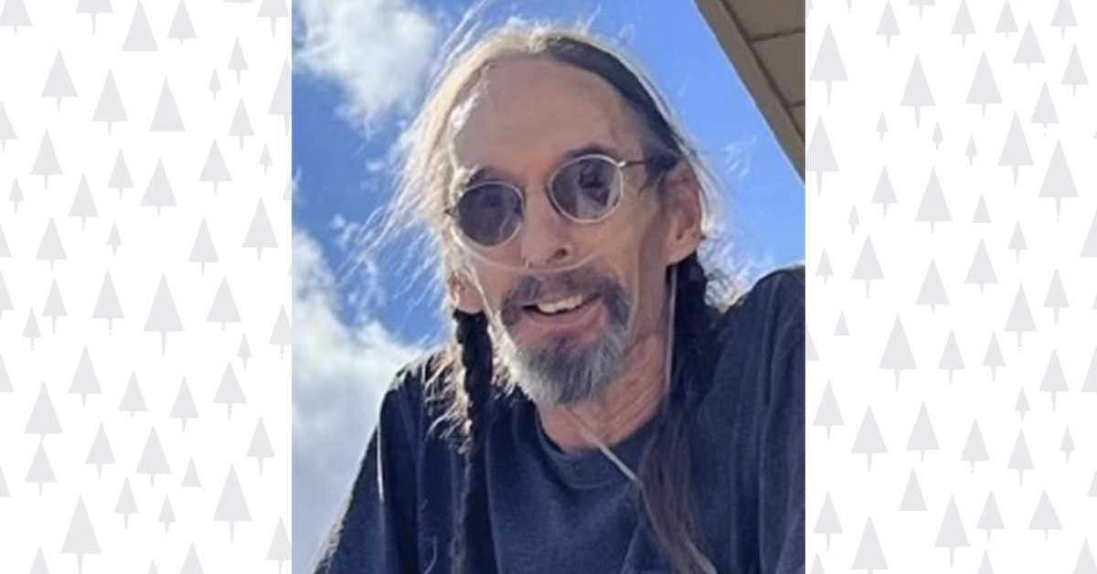 Ray Glenn smiles beneath a blue sky, wearing round sunglasses and braided hair, remembered by the Mt. Hood community as a master craftsman and beloved friend.